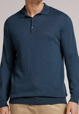 Homme portant un polo à manches longues bleu marine avec deux boutons et un pantalon beige, torse visible, tête partiellement hors du cadre.
