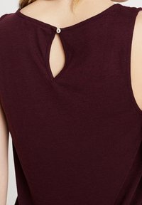 Burgundy ärmlös topp med en nyckelhålsöppning bak, säkrad med en liten vit knapp; mjuk tygkvalitet med en avslappnad passform.