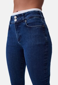 Jeans in denim blu scuro con vita alta, due bottoni argentati e vestibilità aderente. Tessuto liscio con lievi scoloriture sulle cosce.