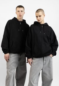 Sorte zip-up hetter i en avslappet passform, med frontlommer og broderte detaljer. Kombinert med lysegrå løstsittende jeans.