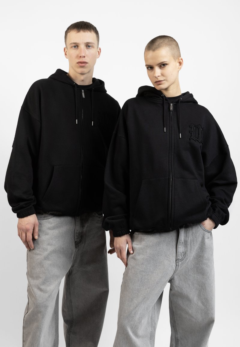 Sorte zip-up hetter i en avslappet passform, med frontlommer og broderte detaljer. Kombinert med lysegrå løstsittende jeans.