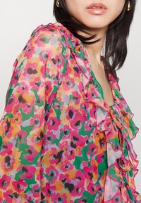 Blouse florale avec des motifs vibrants en rose, vert et orange, dotée de bords volantés et d'un détail noué au niveau du col. Tissu léger.