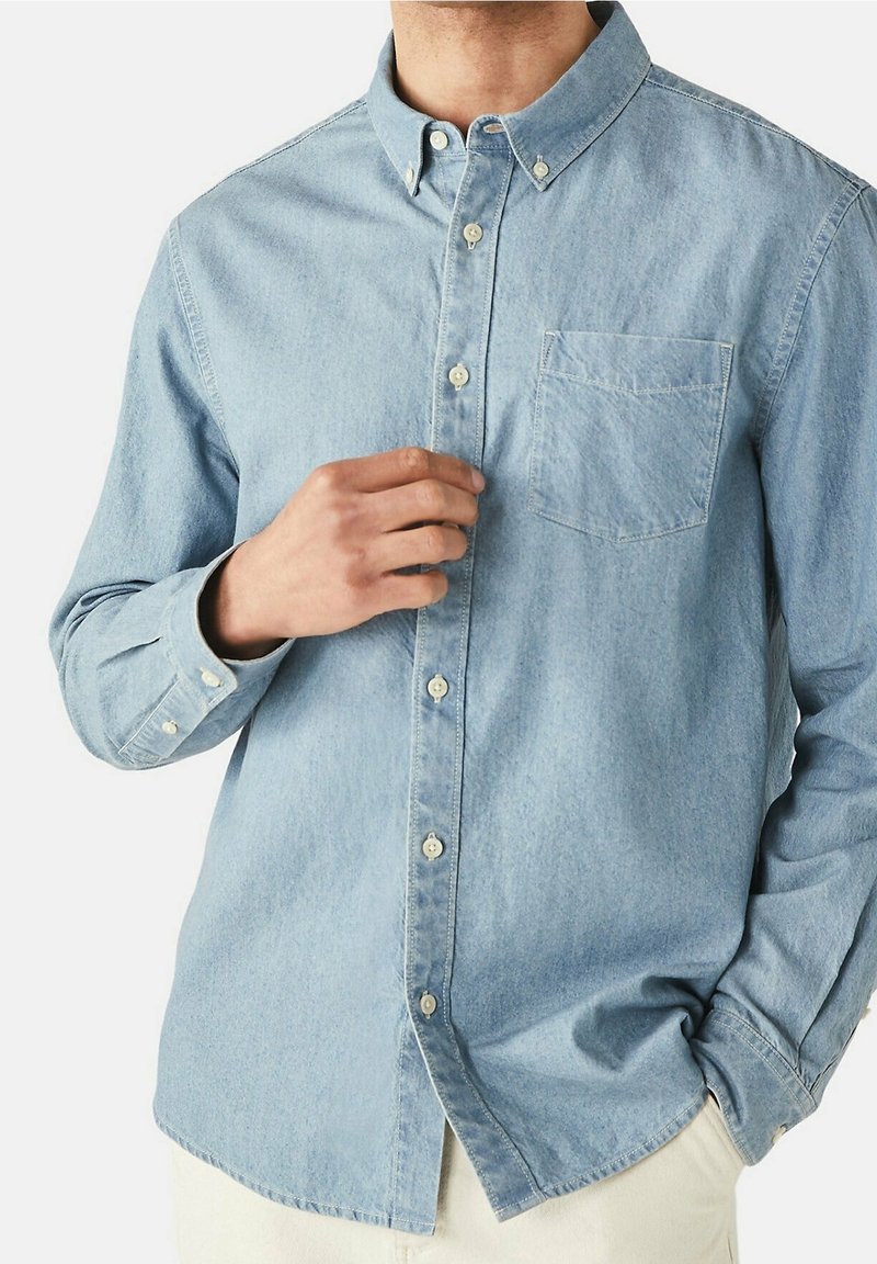 Homme portant une chemise en denim bleu clair avec poche poitrine, une main boutonnant devant, l'autre main dans la poche d’un pantalon beige.