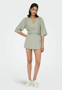 Hellgrünes kurzes Kleid mit V-Ausschnitt, Puffärmeln und Wickeldesign, das über eine seitliche Knopfdetail und eine glatte Textur verfügt.