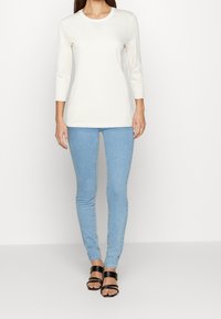 Langärmliges Creme-Oberteil mit dezenten Streifen, kombiniert mit hellblauen Skinny-Jeans und schwarzen Riemchensandalen. Schlichte, figurbetonte Ausführung.