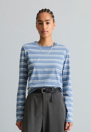 Jeune personne aux cheveux tressés portant une chemise à manches longues rayée bleu et blanc, un pantalon gris, une ceinture noire et un collier en chaîne argentée.