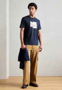 T-shirt de algodão azul-marinho com um design gráfico na frente. Combinado com calças largas bejas e sapatos castanho-escuro. Suéter pendurado no braço.