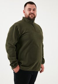 Oliven grøn fleece-pullover med halv lynlås foran, tekstureret stof og detalje ved brystsømmene. Har lange ærmer og en afslappet pasform.