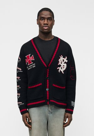 Ung mand iført en sort cardigan med røde kanter, grafiske patches og tekst, kombineret med en sort skjorte og vaskede blå jeans.