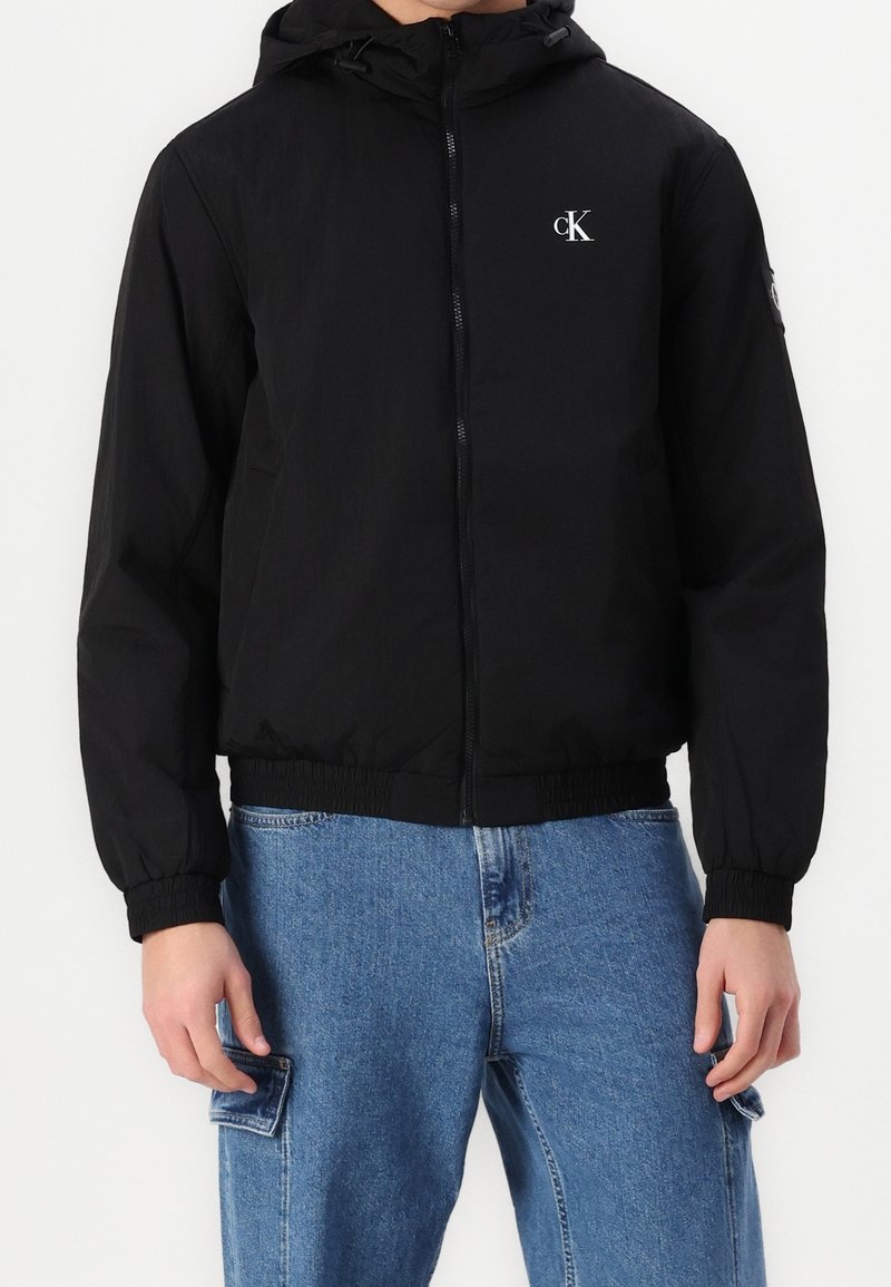 Czarna bluza z kapturem zapinana na zamek, z elastycznymi mankietami i gumką u dołu, z białym logo "cK" na piersi, noszona z niebieskimi jeansami typu cargo.
