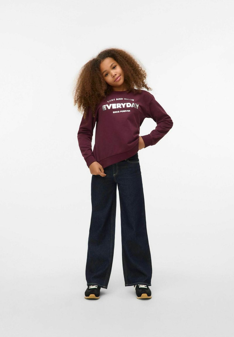 Vinröd hoodie med text, långa ärmar, åtsittande midja. Vida mörk denimjeans. Svarta sneakers med vita och gröna accenter.