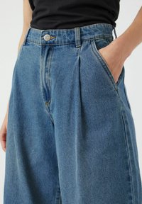 Siniset farkut korkealla vyötäröllä, joissa on laskoksia, metallinen napitus ja sivutaskut. Kangas on sileä.