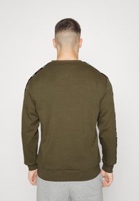 Felpa verde oliva con scollatura tonda e polsini a coste. Presenta dettagli a motivo sulle spalle. Tessuto morbido.