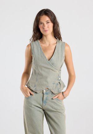 Femme aux longs cheveux foncés porte un haut en denim sans manches croisé et un jean en denim assorti, debout, mains dans les poches, devant un fond uni.