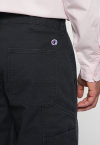 Henkilö, jolla on päällään mustat housut, joissa on neliömäinen takatasku ja Champion-logo vyötärön lähellä, yhdistettynä vaaleanpunaiseen pitkähiharaiseen paitaan.