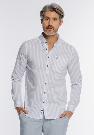 Mężczyzna z siwymi włosami i brodą, ubrany w białą koszulę na guziki z cienkimi pionowymi paskami oraz jasnoszare spodnie, ręce w kieszeniach, neutralny wyraz twarzy.