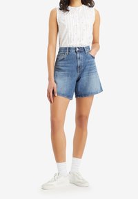 Modré džínové šortky s vysokým pasem a volným střihem, doplněné bílým sleeveless topem s vyšívanými detaily. Celý vzhled dotvářejí bílé tenisky.