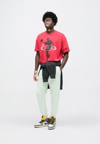 Κόκκινο γραφικό μπλουζάκι με θέμα το μπάσκετ, συνδυασμένο με χακί σκούρο πράσινο παντελόνι φόρμας και μαύρα αθλητικά παπούτσια με κίτρινες λεπτομέρειες.