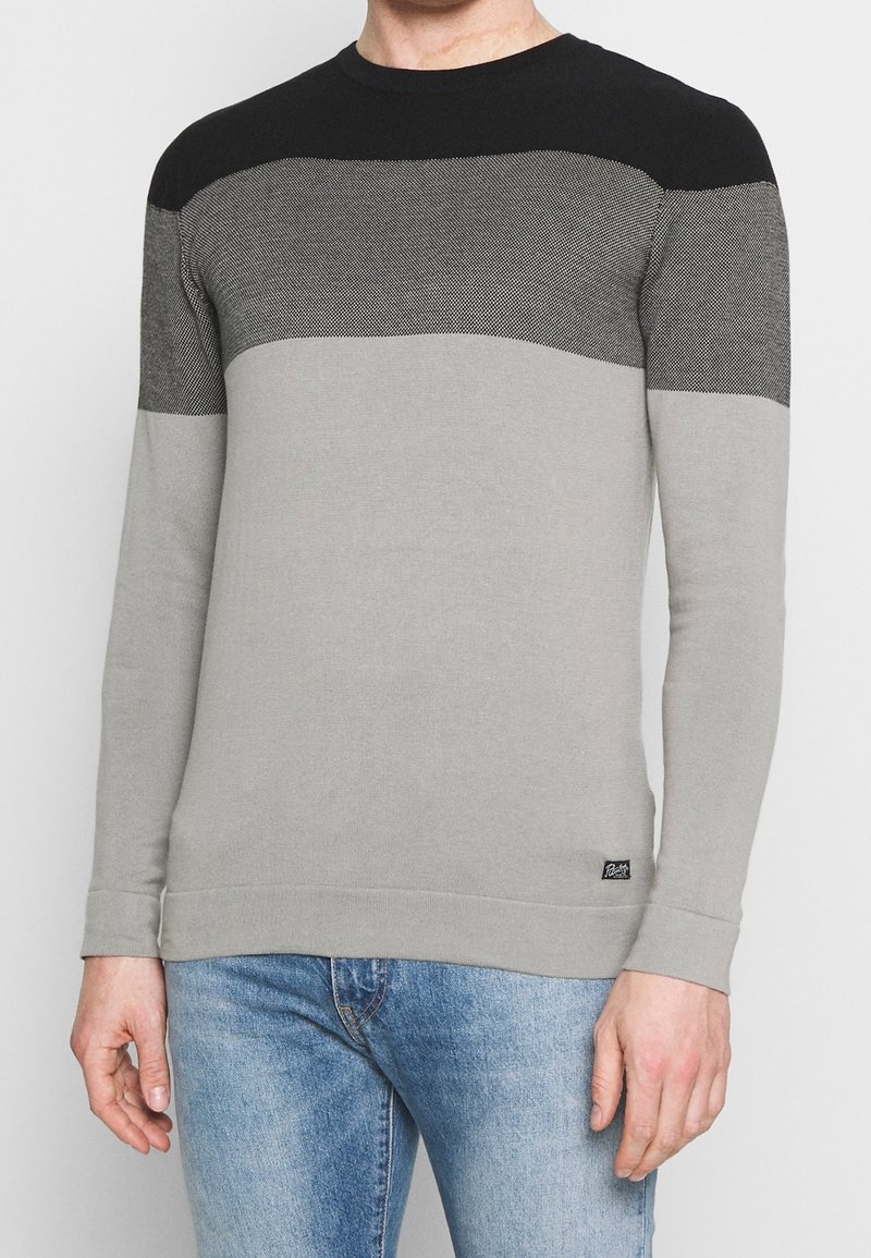 Homme portant un pull à manches longues aux blocs de couleurs horizontaux noir, gris foncé et gris clair, associé à un jean bleu.
