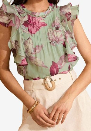 Vrouw die een groene mouwloze blouse met roze bloemenprint en gerimpelde mouwen draagt, gecombineerd met beige hoge taillebroeken en een gouden armband.