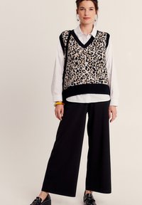 Beige en zwarte gebreide vest met luipaardpatroon, gedragen over een wit shirt. Gecombineerd met ruime zwarte broeken en glanzende zwarte schoenen.