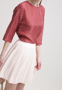 Blusa rossa a fantasia con maniche a tre quarti abbinata a una mini gonna plissettata rosa chiaro. Tessuto liscio con una sottile texture.