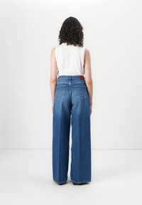 Calças de denim azul de pernas largas com uma cintura alta, apresentando bolsos traseiros e um logo em pele. O tecido tem uma textura clássica desgastada.