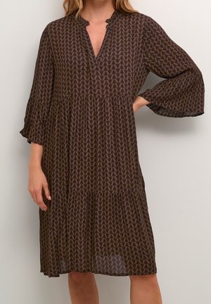 Femme portant une robe marron ample jusqu'aux genoux avec un motif triangulaire géométrique et des manches évasées trois-quarts.