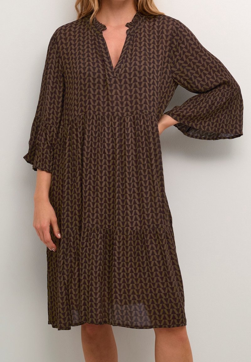 Femme portant une robe marron ample jusqu'aux genoux avec un motif triangulaire géométrique et des manches évasées trois-quarts.