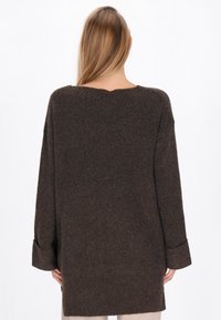 Pulover tricotat lung, maro, cu o croială lejeră, mâneci largi și textură côtée. Dispune de un decolteu adânc la spate pentru un plus de detaliu.