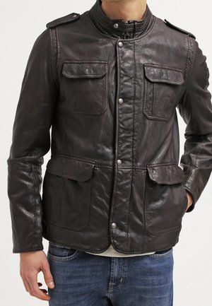 Homme portant une veste en cuir marron foncé avec des poches à rabat à l'avant et des boutons-pression, associé à un jean bleu sur un fond uni.