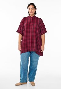 Rood en zwart geruite oversized shirt met korte mouwen en een opstaande kraag, gecombineerd met lichtblauwe rechte jeans en beige gevlochten instappers.