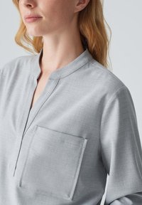 Blusa grigia leggera con scollatura a mezza abbottonatura, dotata di taschino sul petto e vestibilità comoda. Tessuto liscio e design semplice.