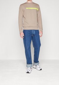 Beige sweatshirt med ett grönt rand som har texten "BOSS". Kombinerad med lösa blå jeans och vita sneakers med grå detaljer.