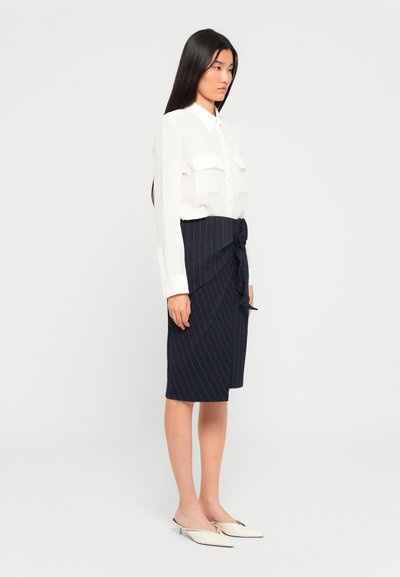 Vrouw met lang donkerhaar staat in een witte blouse met knopen, een marineblauwe strikrok met streepjes en witte puntige lage hakken.
