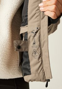 Beige bunda s černým lemováním, vyrobená z recyklované tkaniny. Disponuje vodoodpudivou úpravou, vysokou izolační schopností a technickými ikonami.