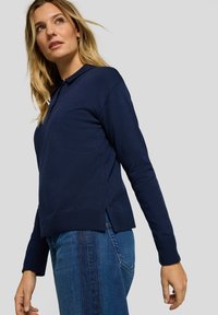 Marineblaues geripptes Pullover-Sweatshirt mit Kragen im Polo-Stil, langen Ärmeln und Schlitzen am Saum, kombiniert mit blauen Jeans.