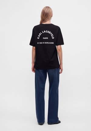 Άτομο με μακριά μαλλιά που φοράει μαύρο t-shirt Karl Lagerfeld Paris με λευκό κείμενο και μπλε παντελόνι με φαρδιά πόδια, στέκεται γυρισμένο από πίσω.