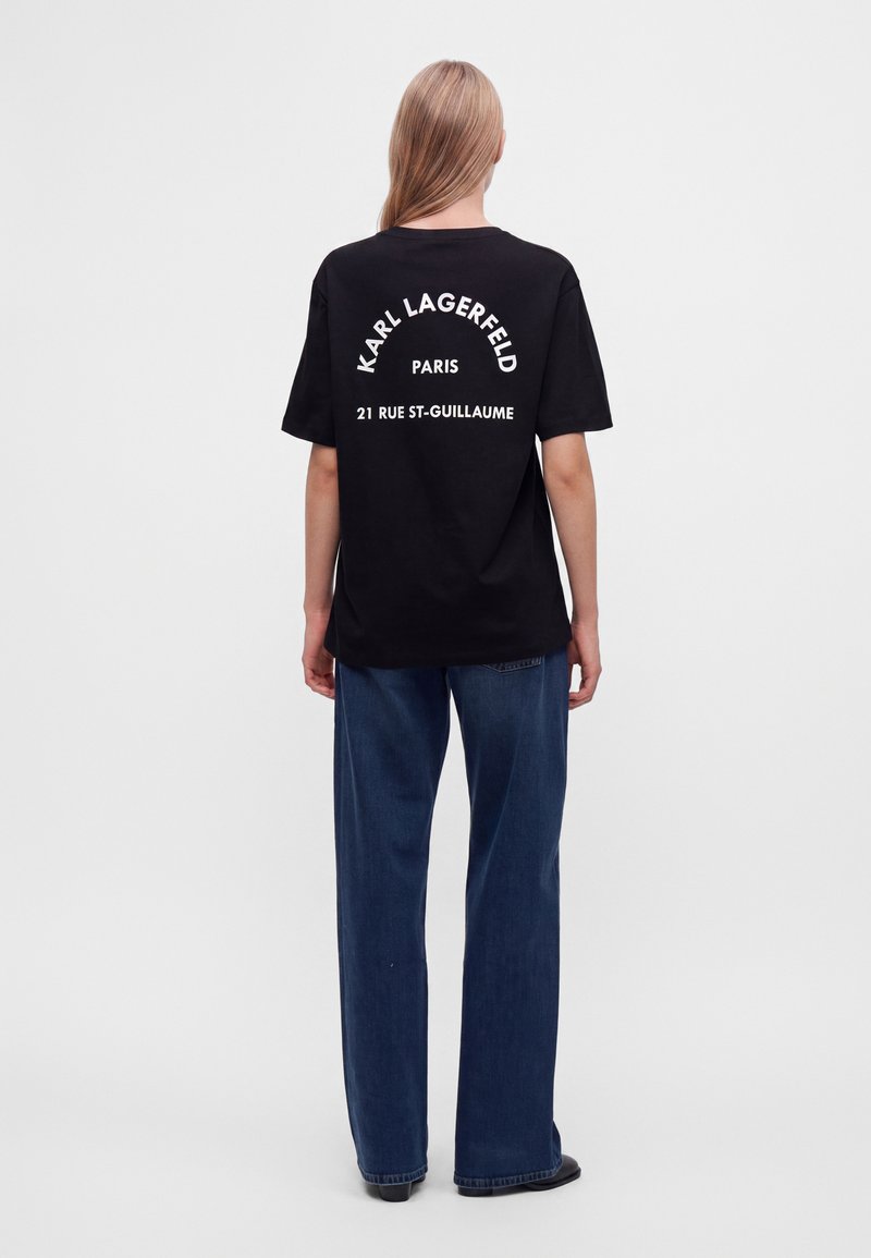 Άτομο με μακριά μαλλιά που φοράει μαύρο t-shirt Karl Lagerfeld Paris με λευκό κείμενο και μπλε παντελόνι με φαρδιά πόδια, στέκεται γυρισμένο από πίσω.