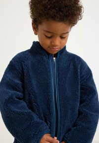 Marineblaue Fleecejacke mit hohem Kragen, Reißverschluss vorne und kontrastierenden Besätzen. Weiches, strukturiertes Material mit weiten Ärmeln und lässiger Passform.