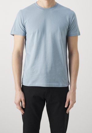 Homme portant un t-shirt bleu clair uni à manches courtes et un pantalon noir, debout devant un fond clair uni.
