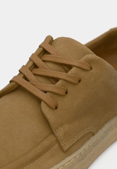 Pantof din piele întoarsă de culoare maro deschis, cu vârf rotund și suprafață texturată. Dispune de șireturi asortate și talpă deschisă la culoare, cu cusături vizibile.
