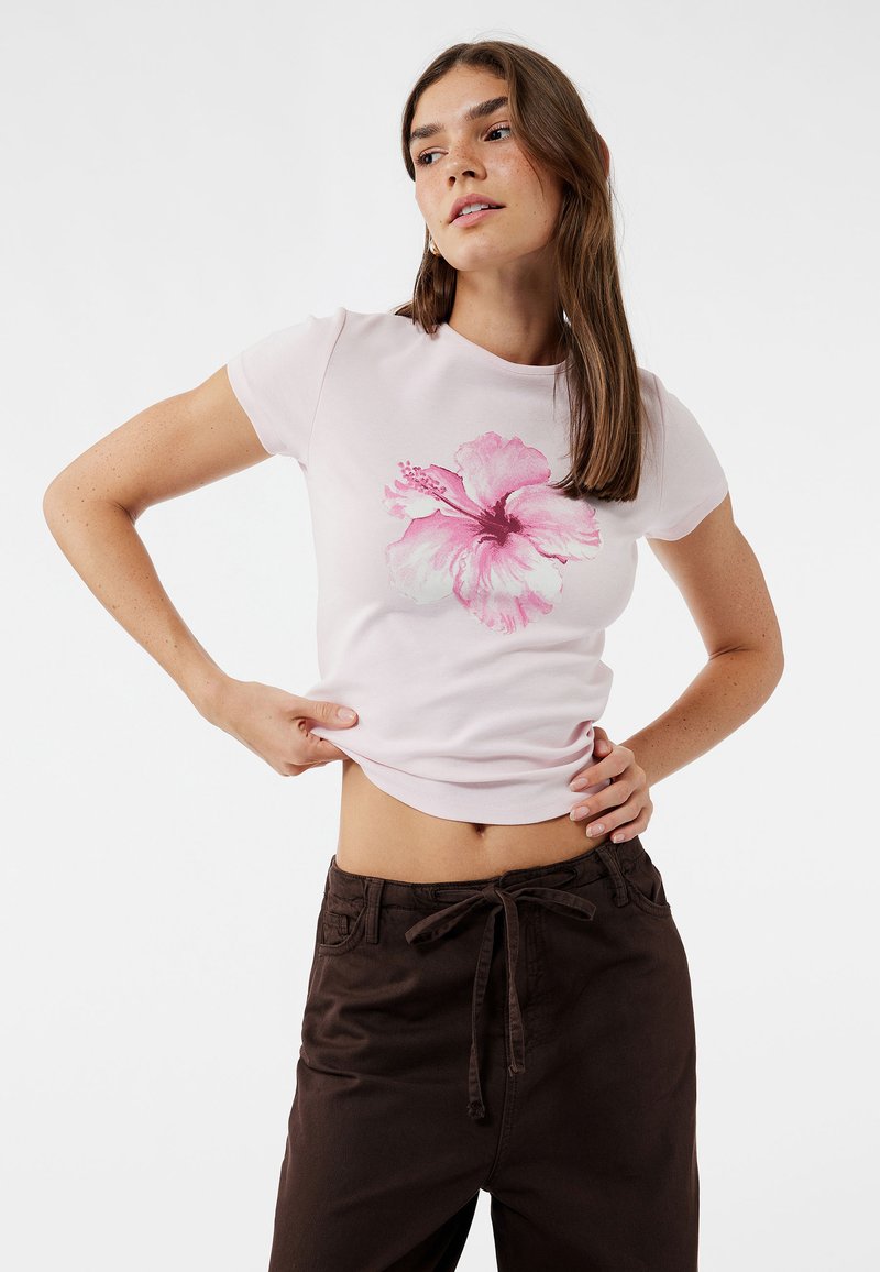 Lys pink fitted t-shirt med et stort print af en pink hibiscusblomst i midten. Bæres med brune højhalset bukser.