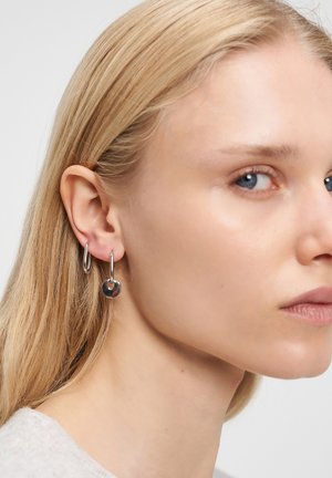 Nahaufnahme eines Ohrs einer Frau, das zwei silberne Creolen zeigt, eine schlicht und eine mit einem kleinen runden quadratischen Anhänger, mit blonden Haaren und heller Haut.