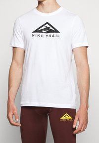 Camiseta blanca de algodón con un logotipo triangular negro que lleva el texto "NIKE TRAIL". Mangas cortas, cuello redondo, combinada con leggings color burdeos.
