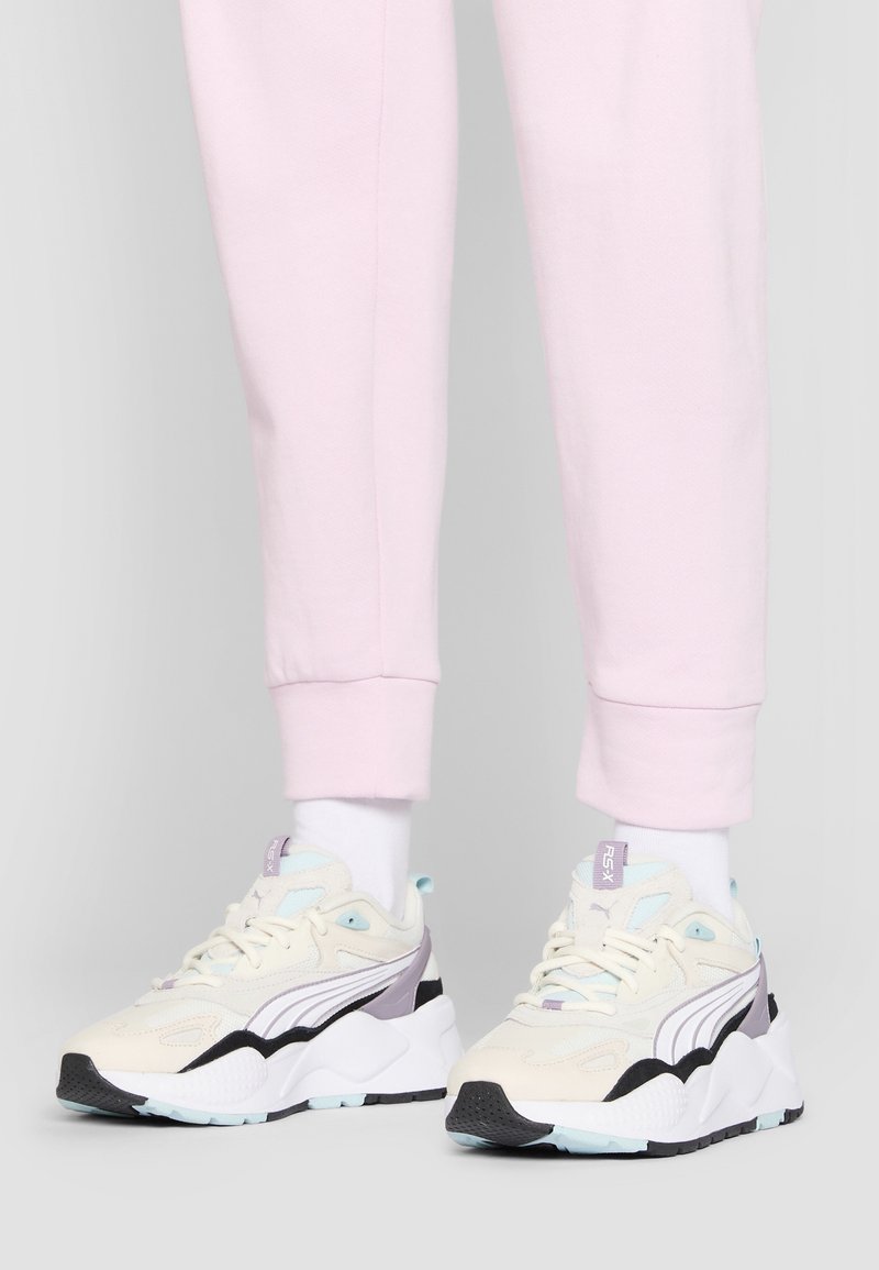 Chaussures de sport en crème, bleu clair et noir. Matériaux en suède et mesh avec une semelle blanche épaisse et des accents violets. Portées avec un pantalon de jogging rose.