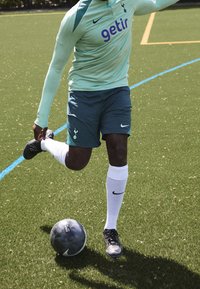 Mintunvihreä pitkähihainen paita, jossa on "getir"-logo, tummanvihreät shortsit, joissa on Spurs-logomerkki, valkoiset sukat ja mustat jalkapallokengät, potkaisee mustaa palloa nurmella.