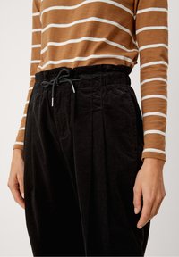 Brun och vit randig långärmad topp inskjuten i svarta dragsnörkordbyxor som bärs av en person som står mot en enkel bakgrund.