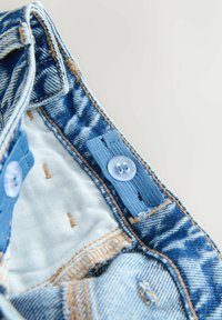 Denim beklædningsdetalje med blåt stof, synlig syning, en lyseblå knap og struktureret indre foring med kontrasterende brune accenter.