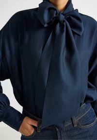 Marineblå bluse med en stor sløjfe ved kraven, lavet af glat stof. Har puffy ærmer og en afslappet pasform. Jeans i denim er synlige.